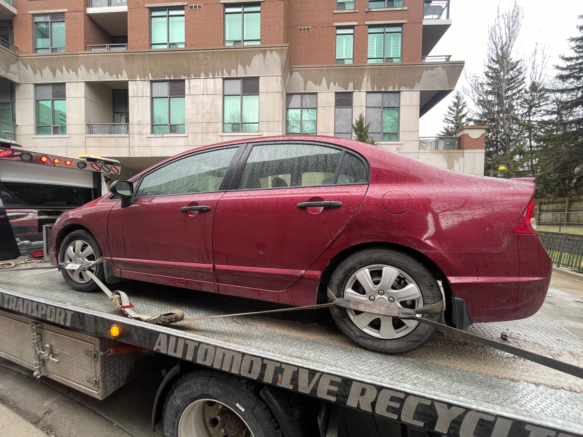 Red sedan secured for pickup outside a condominium building on a rainy day